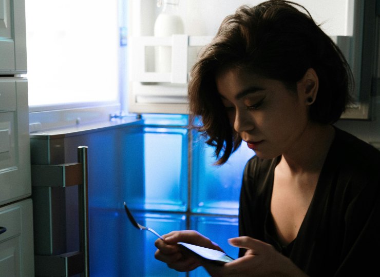 donna al buio seduta accanto al freezer con in mano un cucchiaino e il cellulare
