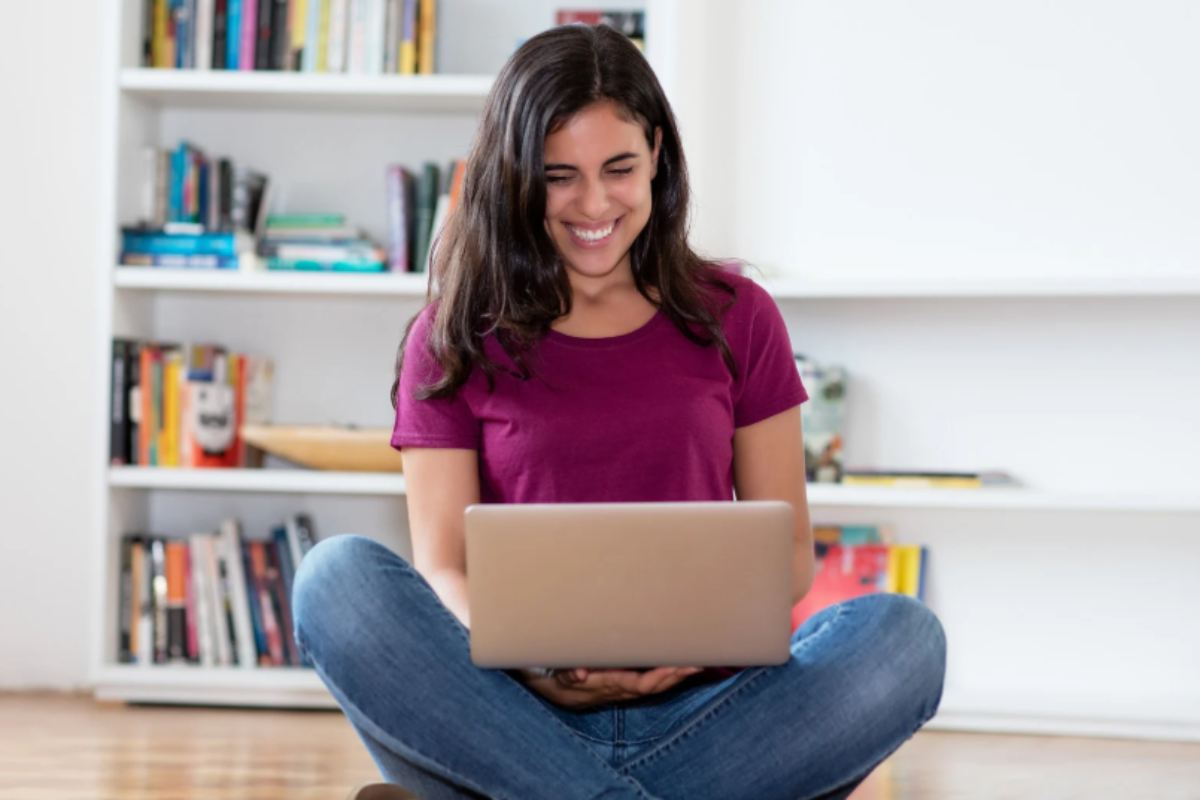 ragazza che sorride seduta sul pavimento con le gambe incrociate e il computer sopra le gambe